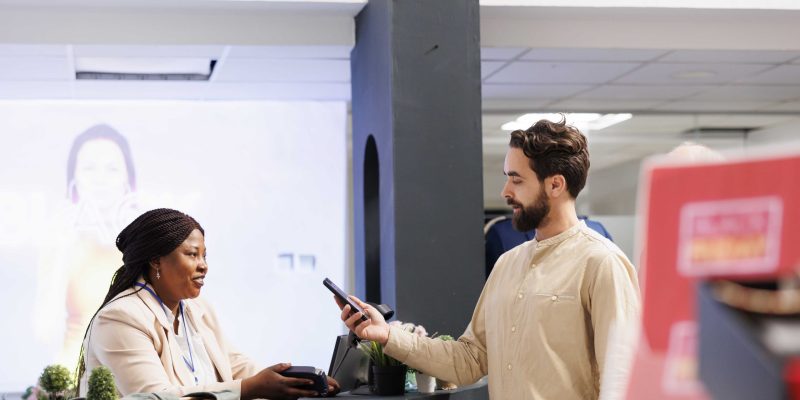 Young man standing at retail checkout counter holding smartphone paying for purchases, using nfc technology to pay for clothes while shopping in clothing store, customer making contactless payment