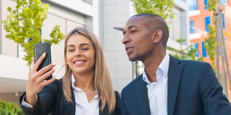 Diverse business colleagues having fun outside. Business man and woman sitting near office building, taking selfie, smiling at phone screen. Selfie concept